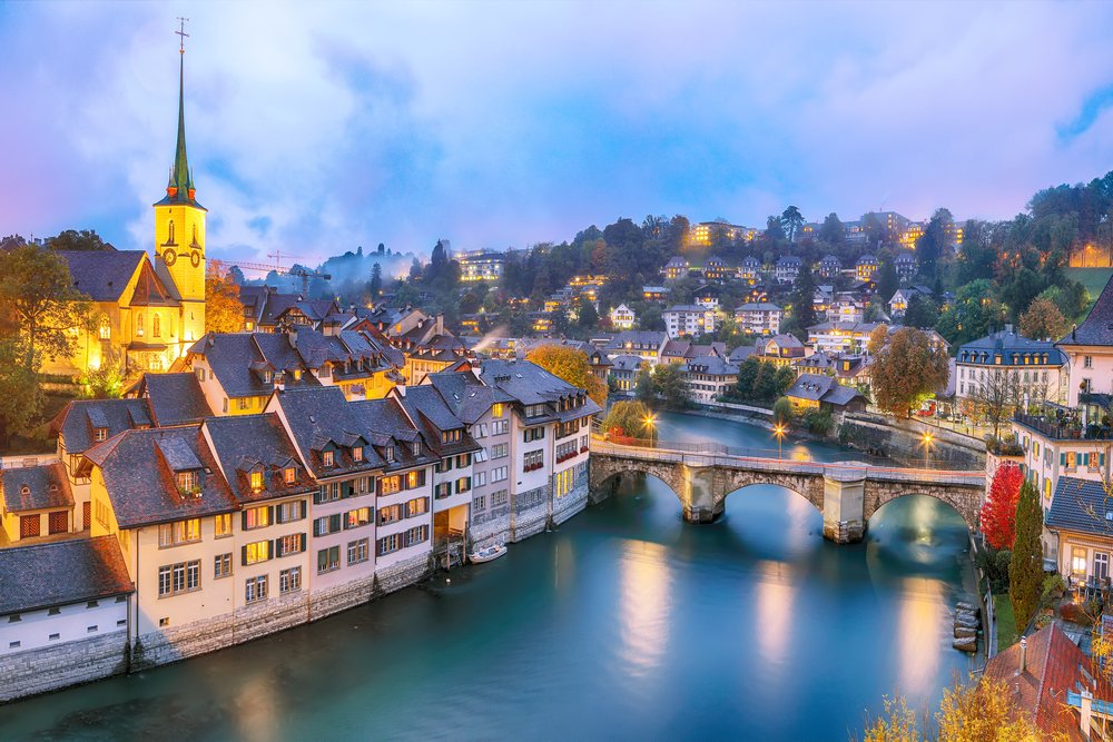 Die Berner Altstadt in der Dämmerung von der Aare aus: die beleuchteten Häuserzeilen am Hang, davor die Brücke und das türkisfarbene Wasser.