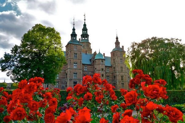 Schloss Rosenborg hinter einem Beet leuchtend roter Rosen, daneben eine Trauerweide; darüber ein bewölkter Himmel.