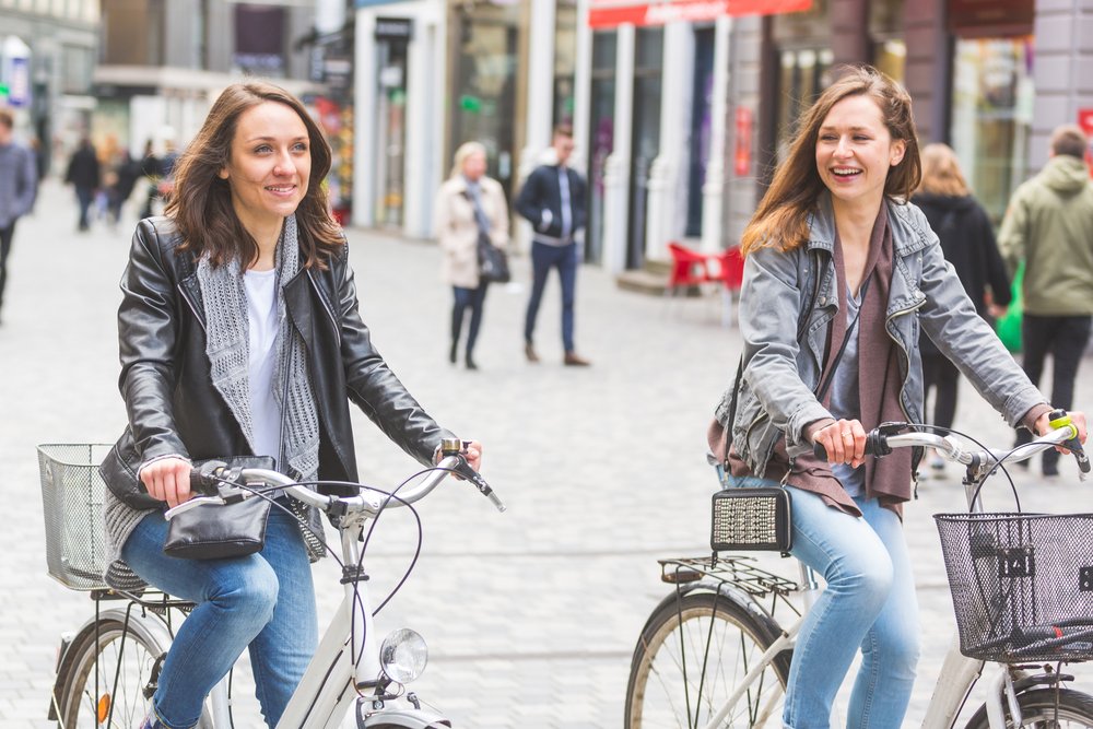Zwei Frauen fahren nebeneinander auf Stadtvelos durch eine belebte Fussgängerzone und unterhalten sich.