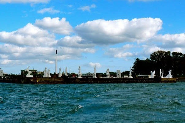 Blick über bewegtes Hafenwasser auf eine flache Uferpromenade mit weissen Schirmen und Bäumen.