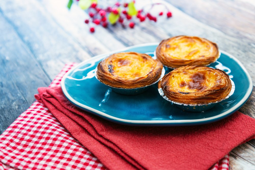 Drei Pastéis de Nata auf einem türkisfarbenen Teller mit gebräunter Oberfläche, daneben eine rot-weiss karierte Serviette.
