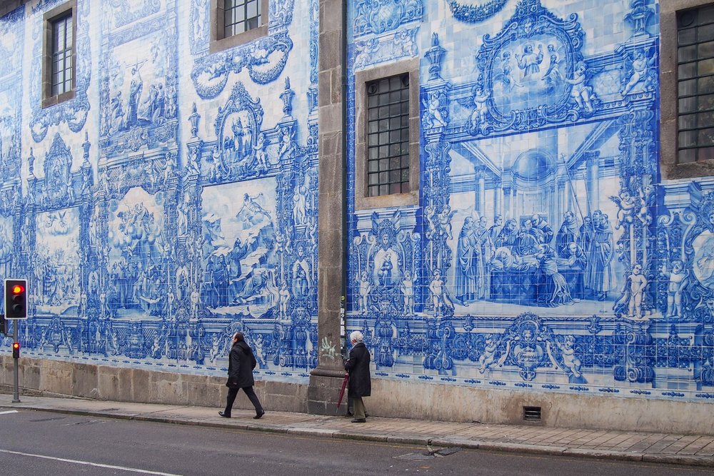 Eine ganze Hauswand aus blau-weissen Azulejo-Kacheln mit gemalten Szenen; zwei Menschen gehen auf dem Gehsteig daran vorbei.