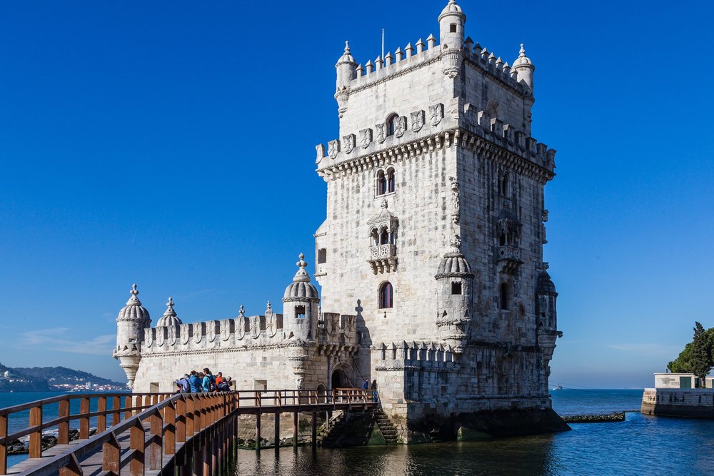 Der Torre de Belém aus hellem Kalkstein im Wasser des Tejo, ein hölzerner Steg führt hinüber, darüber wolkenloser blauer Himmel.