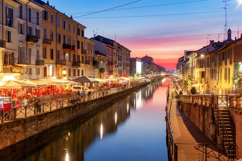 Der Naviglio-Kanal am Abend: Restauranttische unter Schirmen an beiden Ufern, die Lichter spiegeln sich im stillen Wasser.