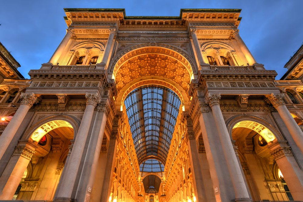 Der Eingangsbogen der Galleria Vittorio Emanuele II zur blauen Stunde, warm beleuchtet, dahinter das Glastonnendach.