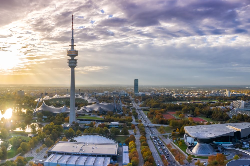 Blick von oben über den Olympiapark: weite Wiesen, Baumgruppen und ein See, dahinter der Olympiaturm und die Stadt im Gegenlicht.