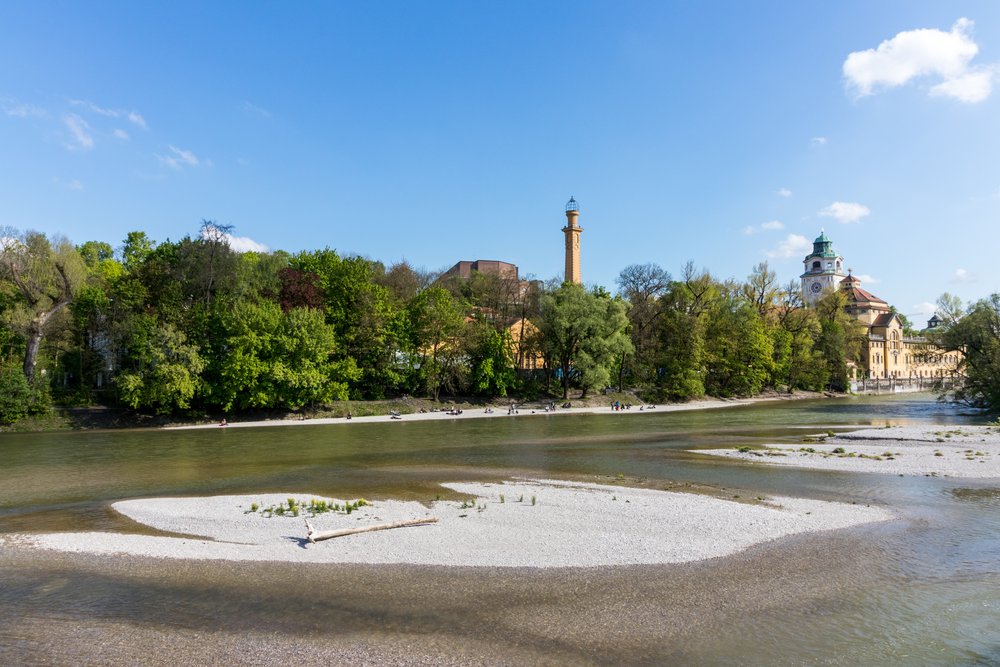 Die Isar in München: eine helle Kiesbank im flachen Wasser, dahinter Bäume und der Turm des Müllerschen Volksbads.