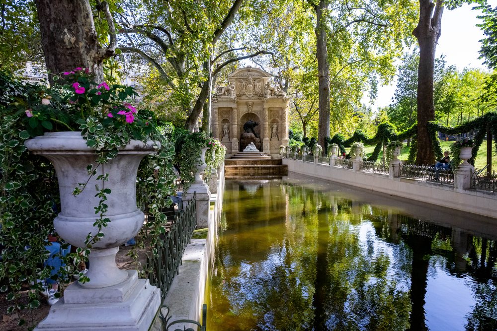 Der Medici-Brunnen im Jardin du Luxembourg: ein langes stilles Wasserbecken zwischen alten Bäumen, Blumenkübel am Rand, Stühle im Schatten.