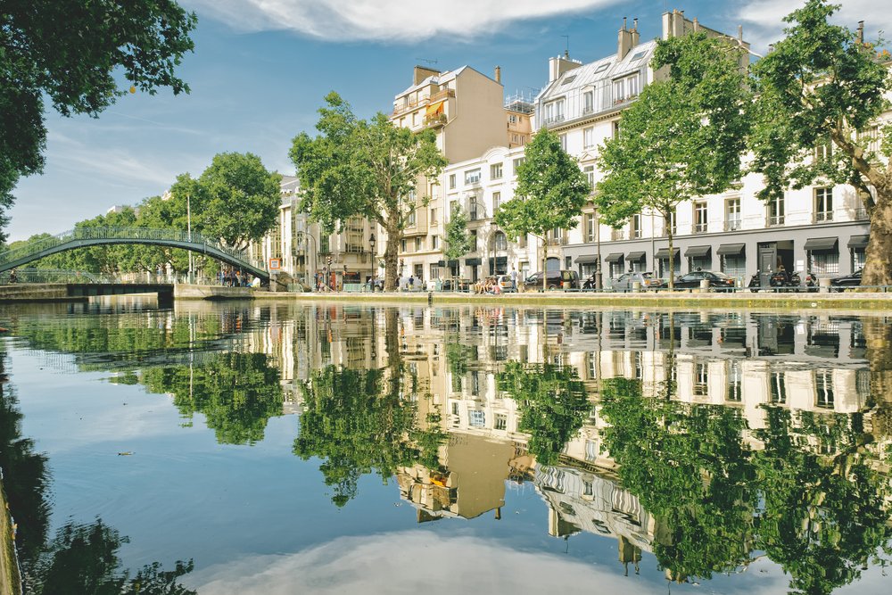 Der Canal Saint-Martin in Paris: eine Fussgängerbrücke über stilles Wasser, in dem sich Platanen und Häuserfronten spiegeln.