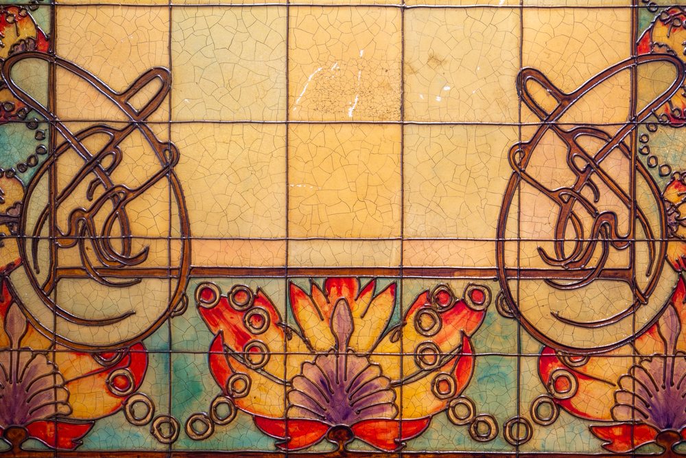 Jugendstil-Kacheln in Ocker, Rot und Türkis: geschwungene Linien, stilisierte Blüten, feine Craquelé-Risse in der Glasur.