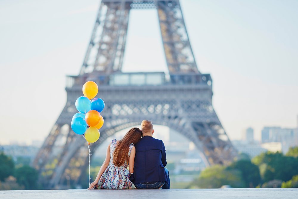 Ein Paar sitzt von hinten gesehen auf einer Mauer, ein Bündel blauer und gelber Luftballons daneben; im weichen Hintergrund der Eiffelturm.