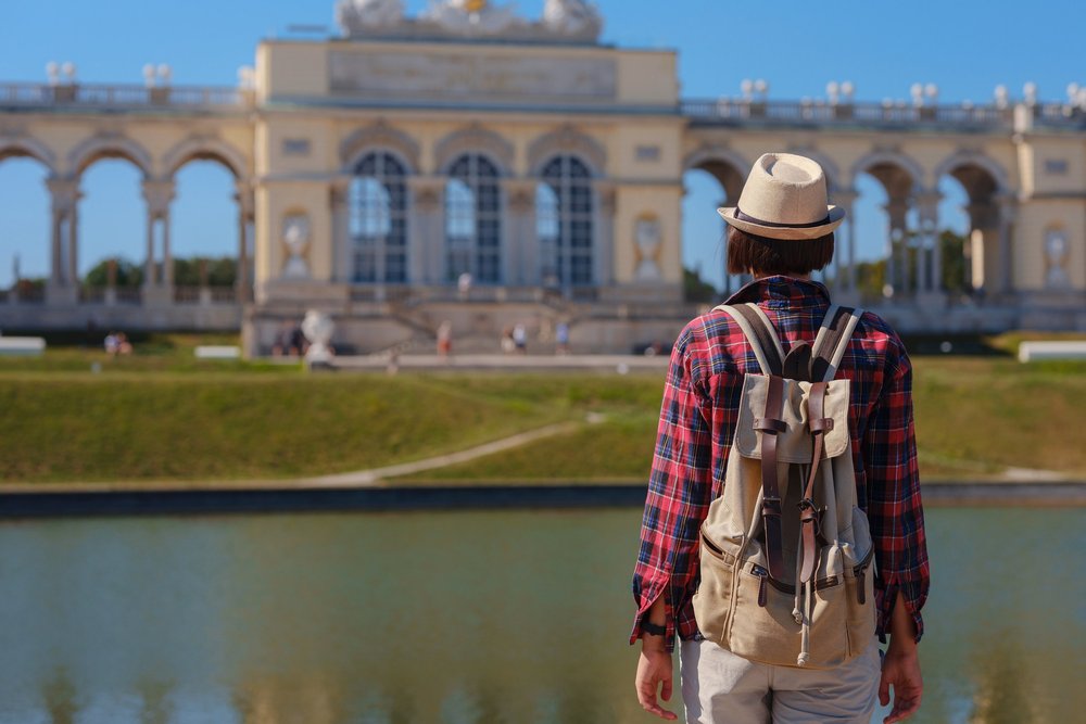 Die Gloriette im Schlosspark Schönbrunn hinter einem stillen Wasserbecken; im Vordergrund geht ein Mensch mit Hut und Rucksack am Ufer entlang.