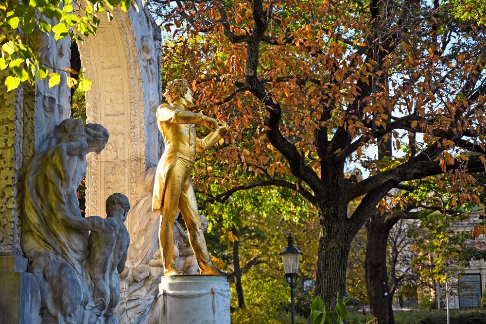 Das vergoldete Johann-Strauss-Denkmal im Wiener Stadtpark, eingerahmt von einem steinernen Bogen und herbstlich gefärbten Bäumen.