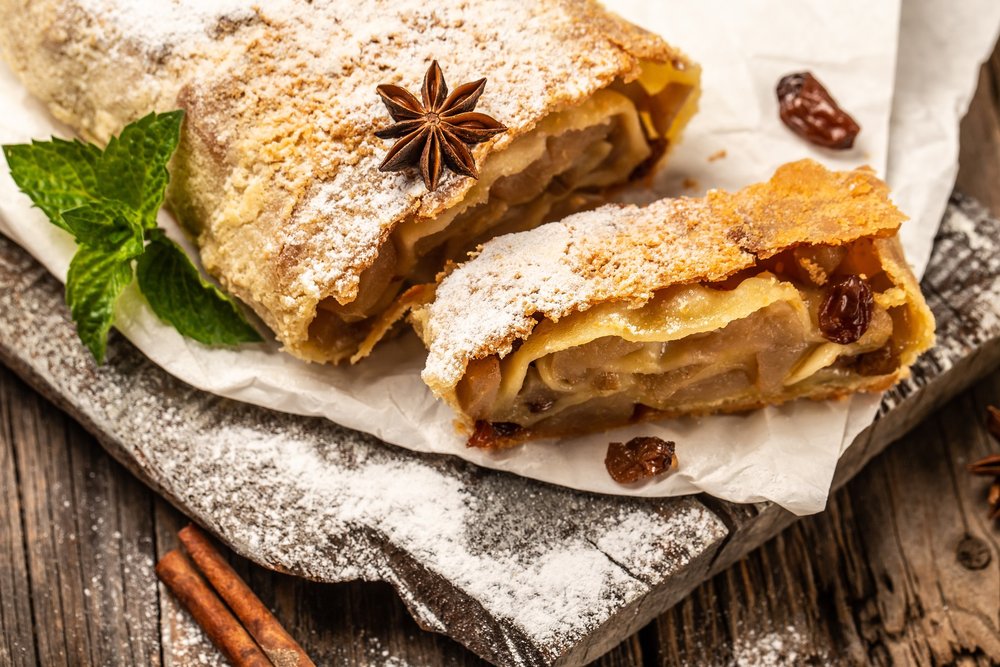 Zwei Stücke Apfelstrudel mit Puderzucker auf Papier, daneben Zimtstangen, ein Sternanis und ein Minzblatt.