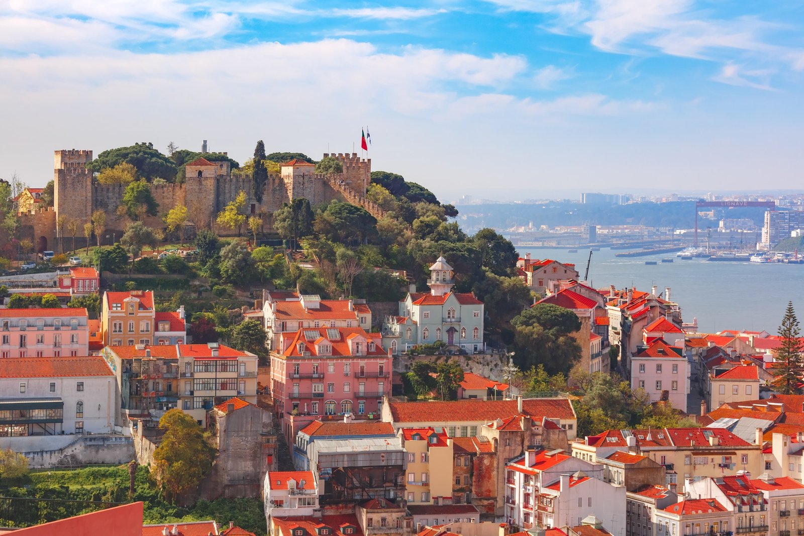 Blick über die Dächer von Lissabon: das Castelo de São Jorge auf dem Hügel, darunter die ockerfarbenen Ziegeldächer der Alfama, dahinter der weite Tejo im Dunst.