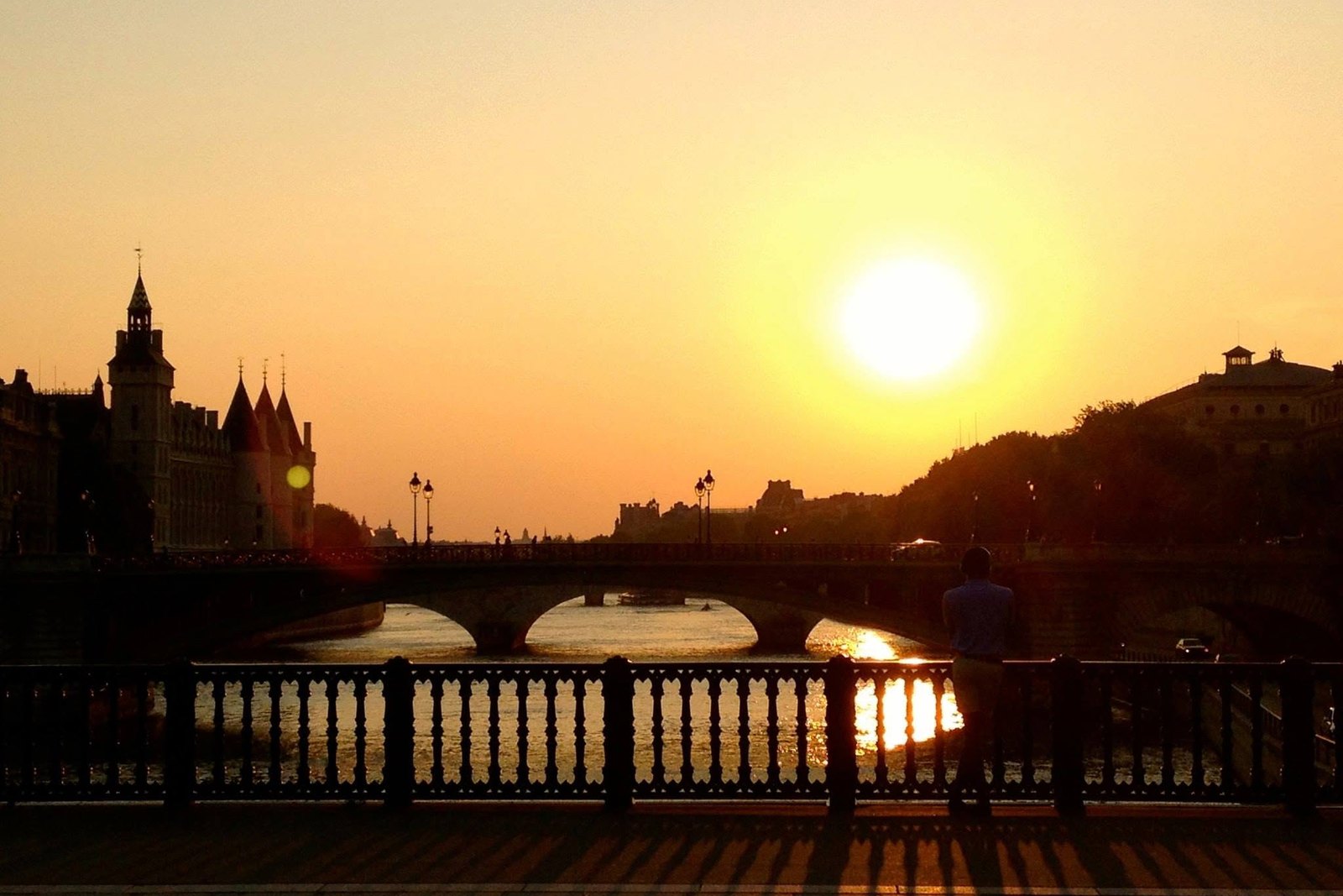 Die Seine bei Sonnenuntergang: die Sonne steht tief über dem Wasser, links die Türme der Conciergerie, in der Mitte eine Brücke, im Vordergrund ein Mensch am Geländer, von hinten gesehen.