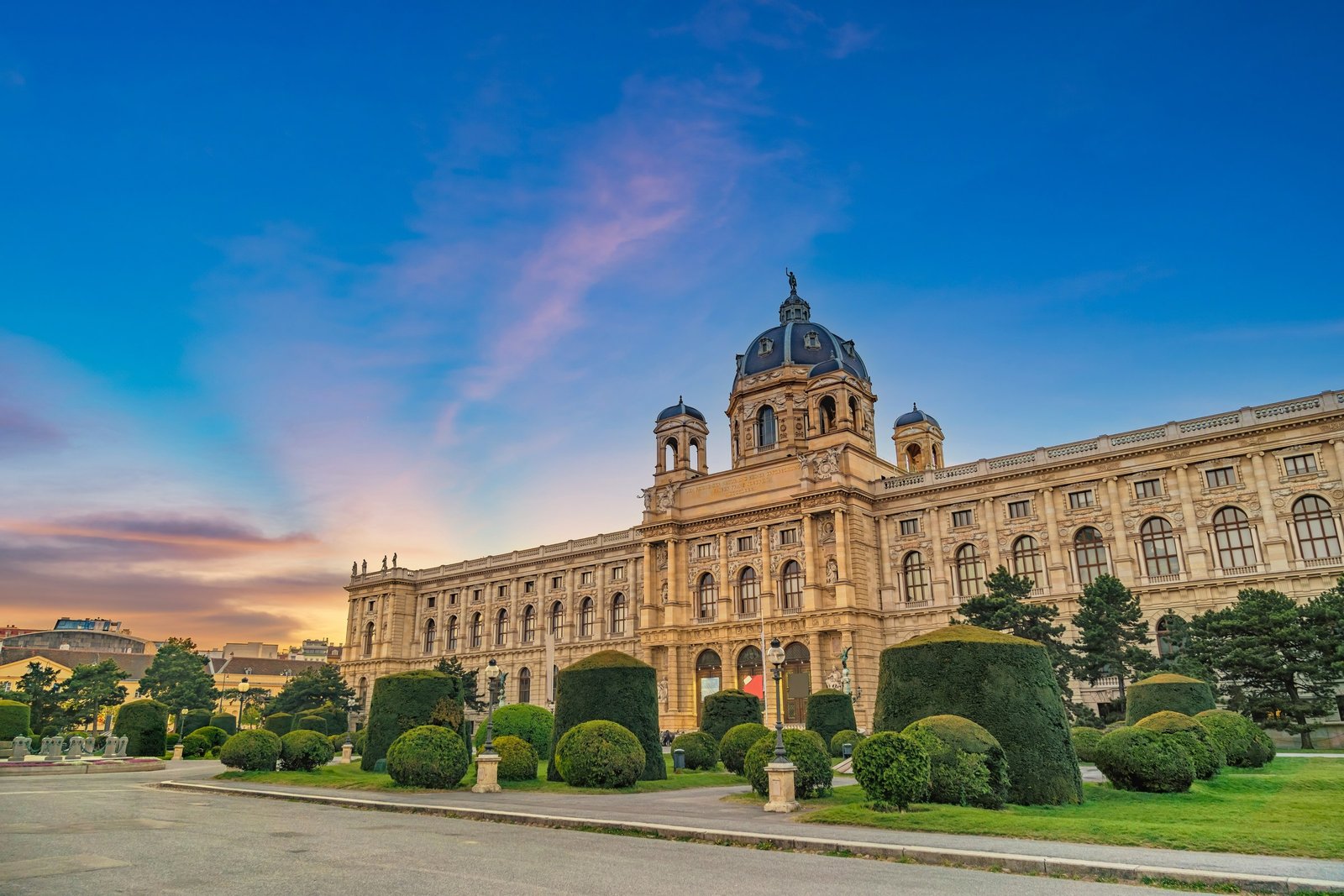 Das Kunsthistorische Museum in Wien in der Abenddämmerung: die breite Fassade und die Kuppel vor einem violett und orange gefärbten Himmel, davor geschnittene Hecken und ein leerer Weg.